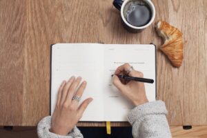 Op deze foto zie je een persoon die haar rolmodellen in een notitieboek aan het schrijven is. Naast haar ligt een bakje koffie en een croissantje.